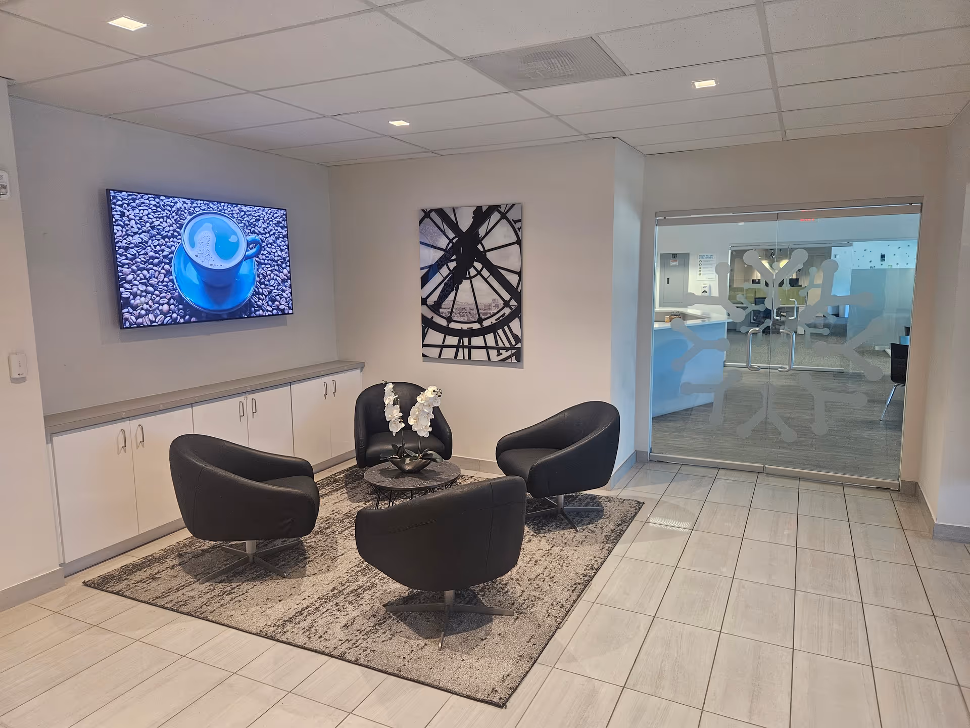 Modern waiting area with four black chairs, glass door, wall art, and a TV.
