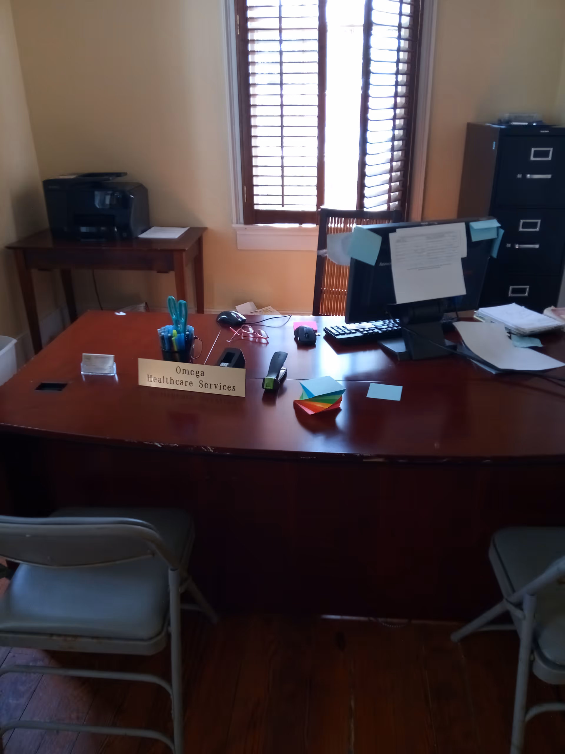 Office desk with computer, printer, and Omega Healthcare Services sign in a quiet workspace.