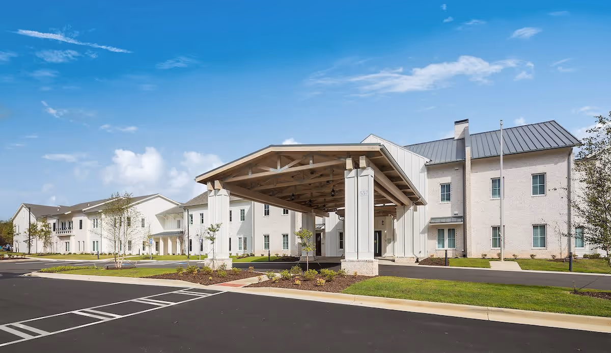 Exterior view of The Claiborne at Shoe Creek senior living facility showing a large covered entrance with wooden beams, white brick buildings, a paved driveway, and a clear blue sky.