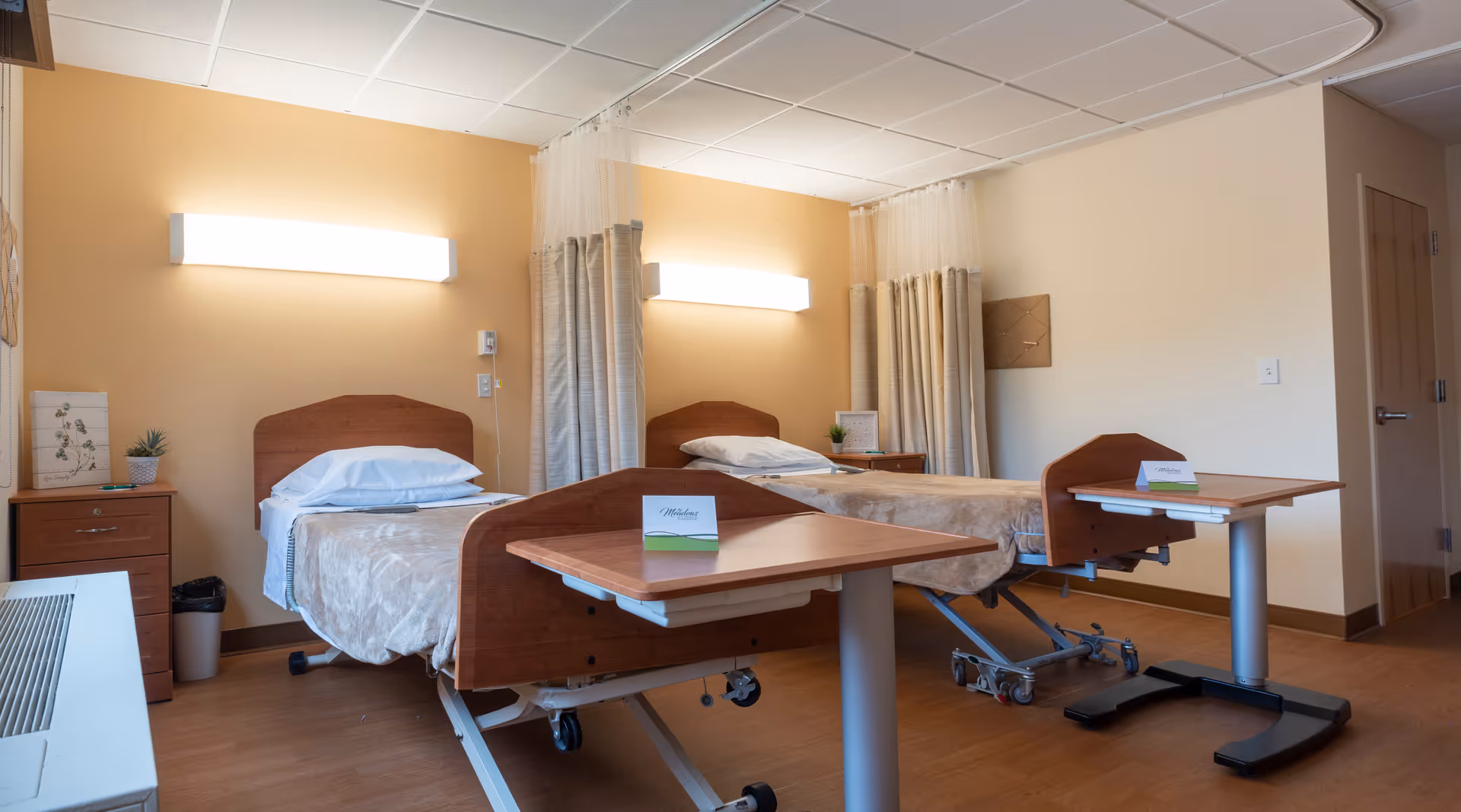 Two adjustable beds with privacy curtains in a furnished senior living patient room.