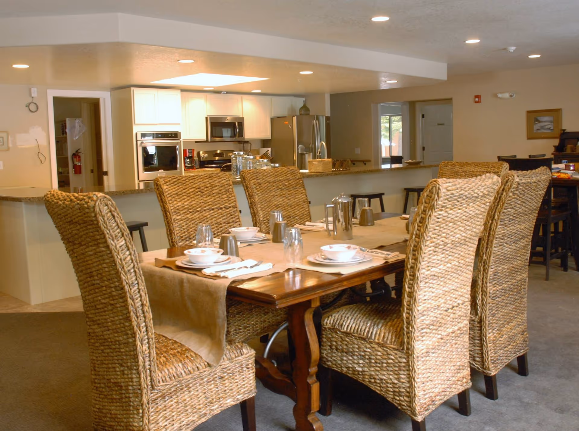 Dining room scene with a long wooden table, wicker chairs, and a kitchen in the background.