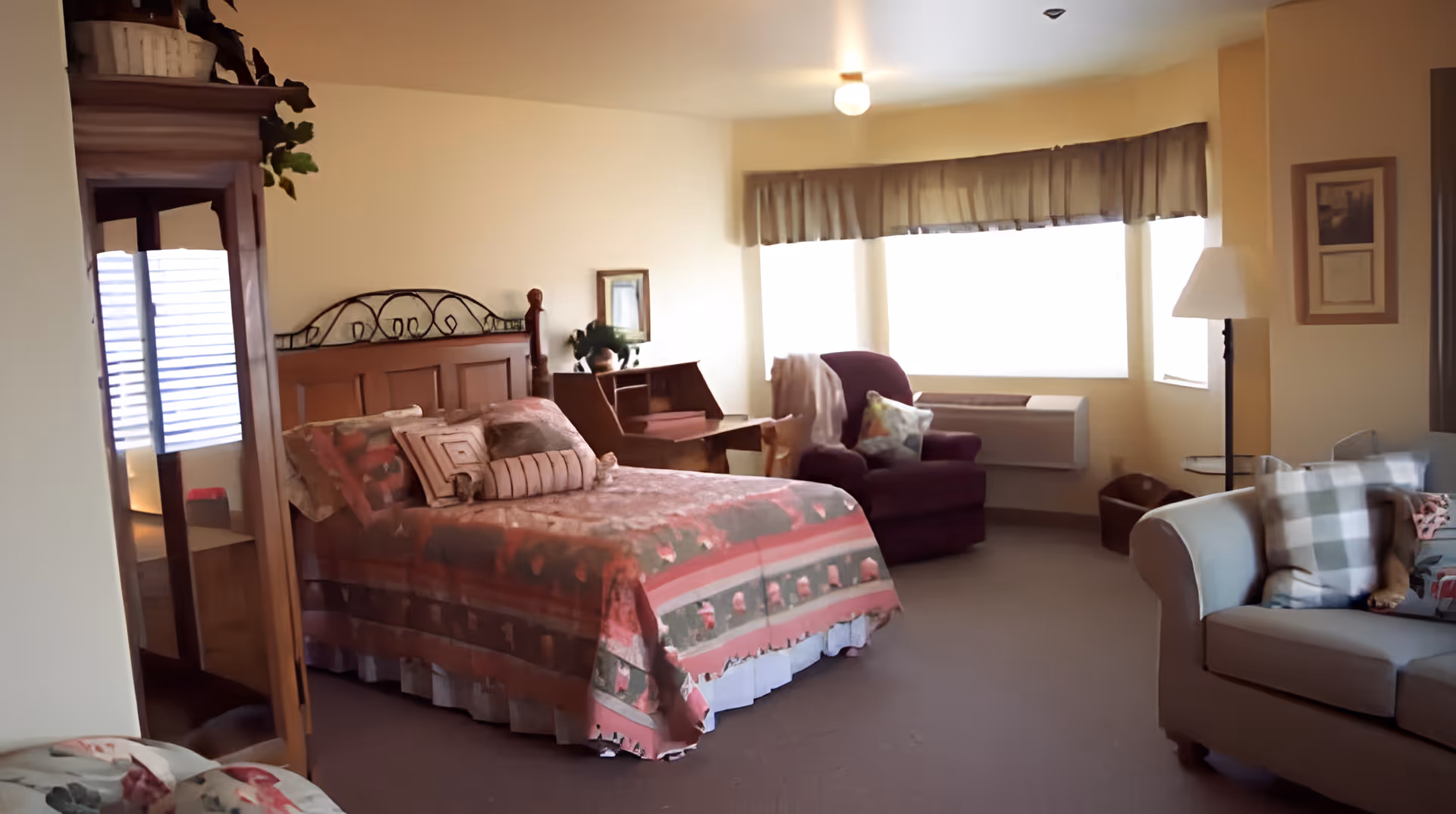 Furnished senior bedroom with a bed, armchair, sofa and a large bay window letting in light.