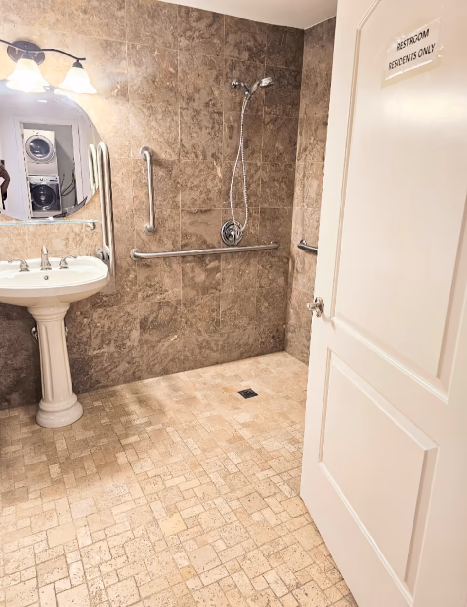 Accessible bathroom with grab bars, pedestal sink, shower head, and tiled walls.