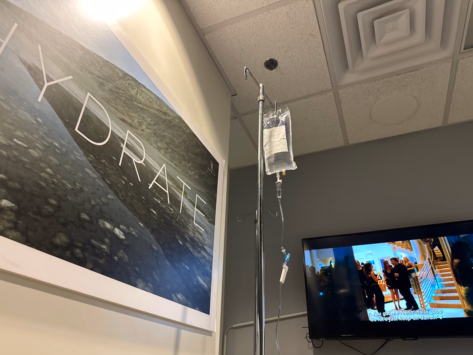IV therapy drip stand with bag in a clinical room beside artwork and a TV.