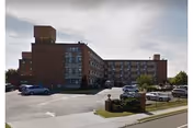 Brick multi-story nursing home building with a driveway, parked cars, and landscaped entrance under a cloudy sky.