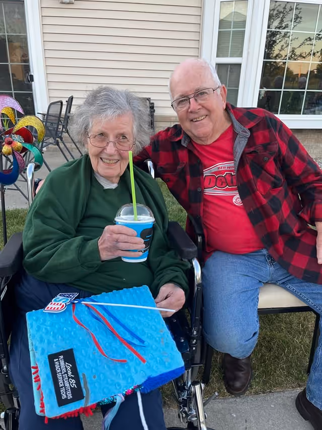 An elderly woman in a wheelchair holding a blue drink with a green straw, sitting next to an elderly man in a red and black checkered shirt and jeans. They are outdoors in front of a beige building with windows, smiling at the camera. The woman has a blue textured blanket with red and blue ribbons on her lap.