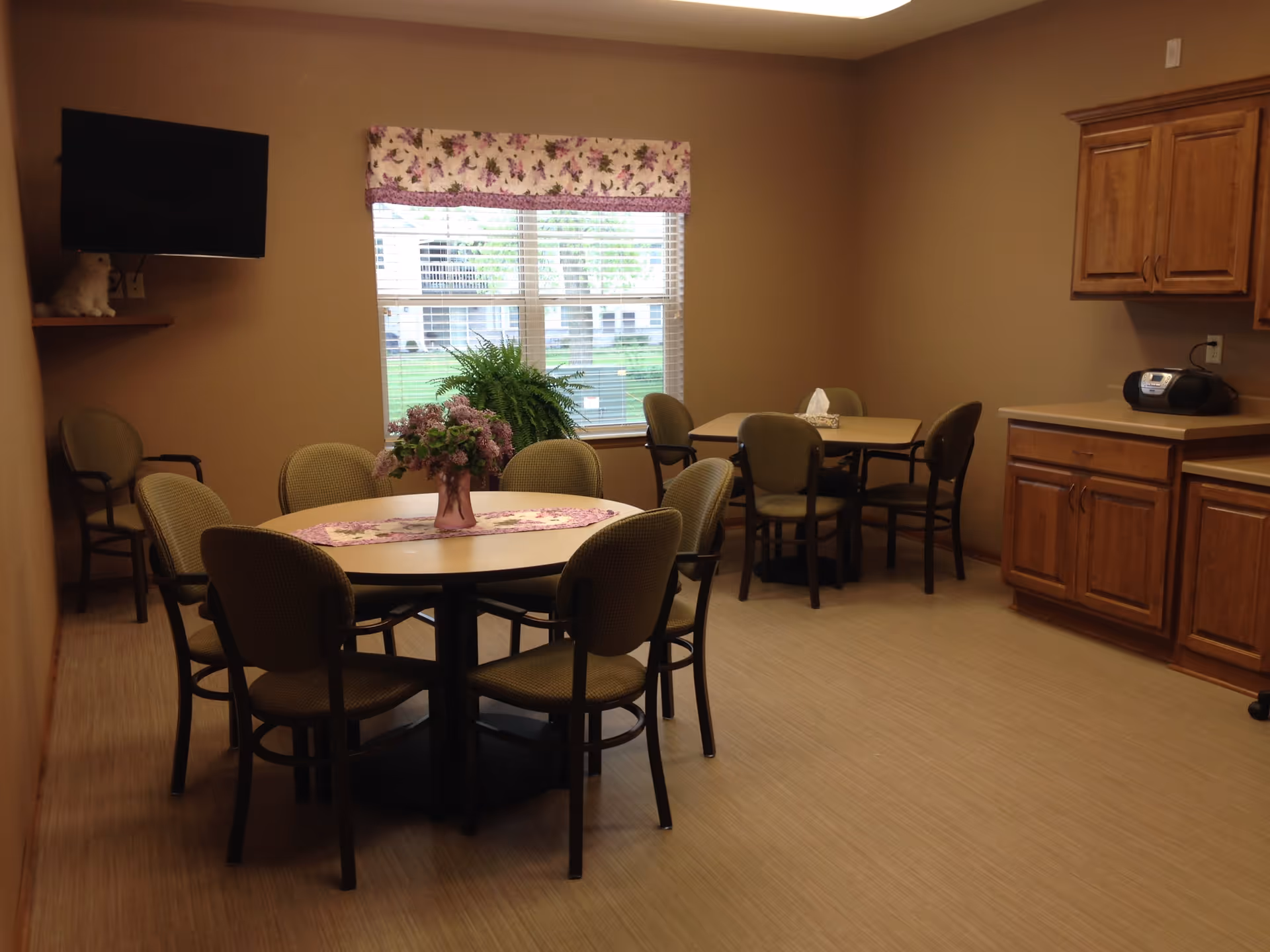 A dining room with two tables surrounded by chairs. One round table has a floral centerpiece and a matching table runner. The room has beige walls, a window with floral valance, wooden cabinets, and a wall-mounted TV.