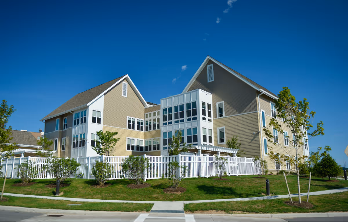 Exterior view of a modern three-story residential building with beige siding and white trim, surrounded by a white fence and landscaped with small trees and shrubs under a clear blue sky.