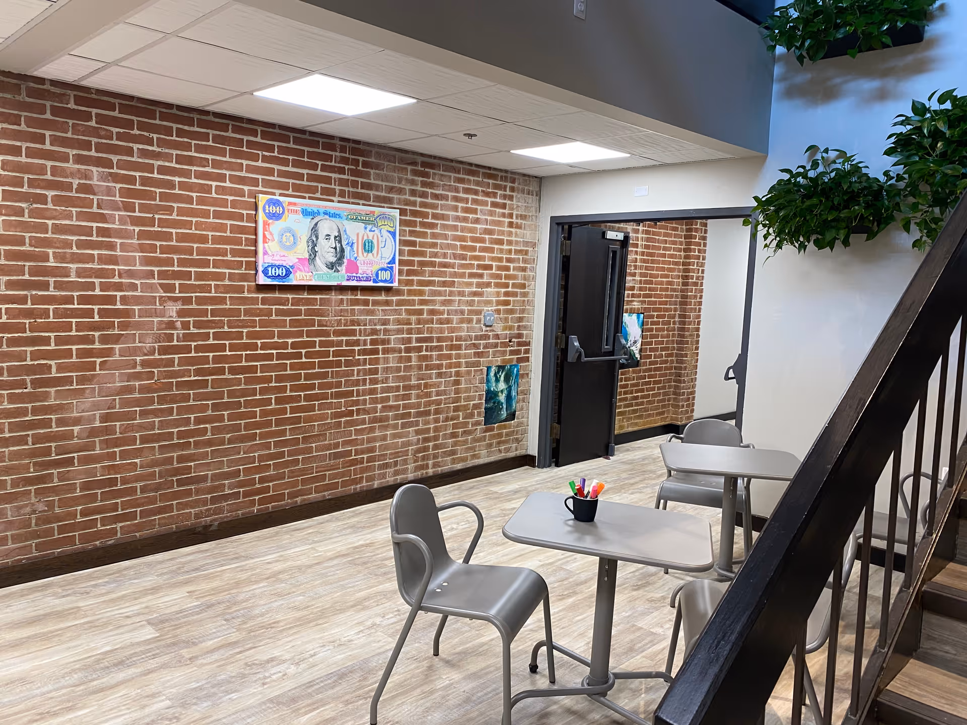 Brick-walled indoor lounge with small tables, chairs, staircase, and potted plants.