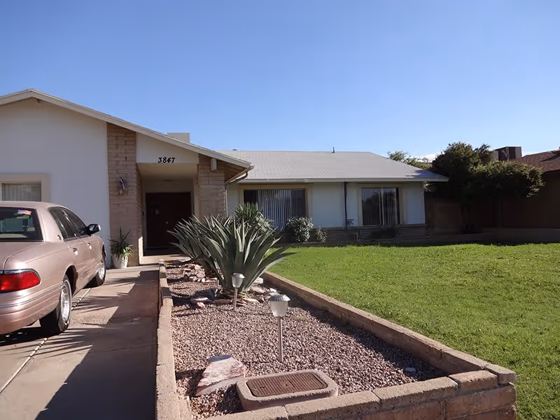 Front exterior view of a single-story residential building with a driveway on the left side where a car is parked. The house has a light-colored facade with a small covered entrance. In front of the house is a landscaped area with gravel, plants, and small garden lights, and a green lawn to the right.