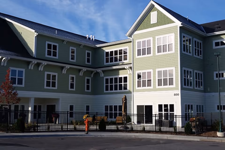 Green multi-story senior living facility exterior with white trim and fenced outdoor seating.