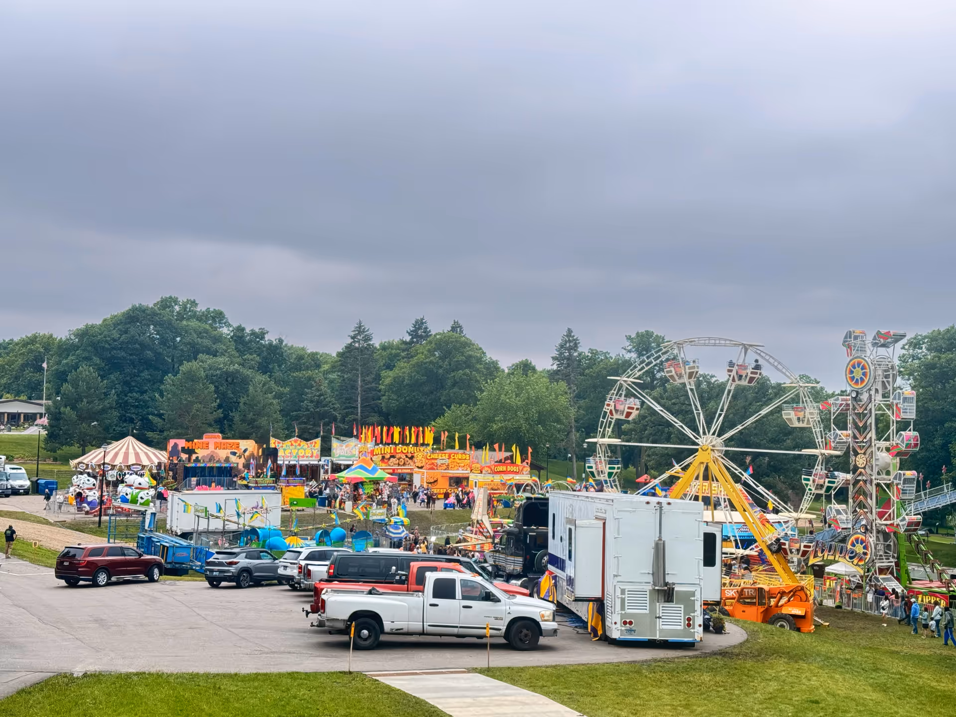 Outdoor carnival with a Ferris wheel, rides, tents, and trucks on a grassy fairground.