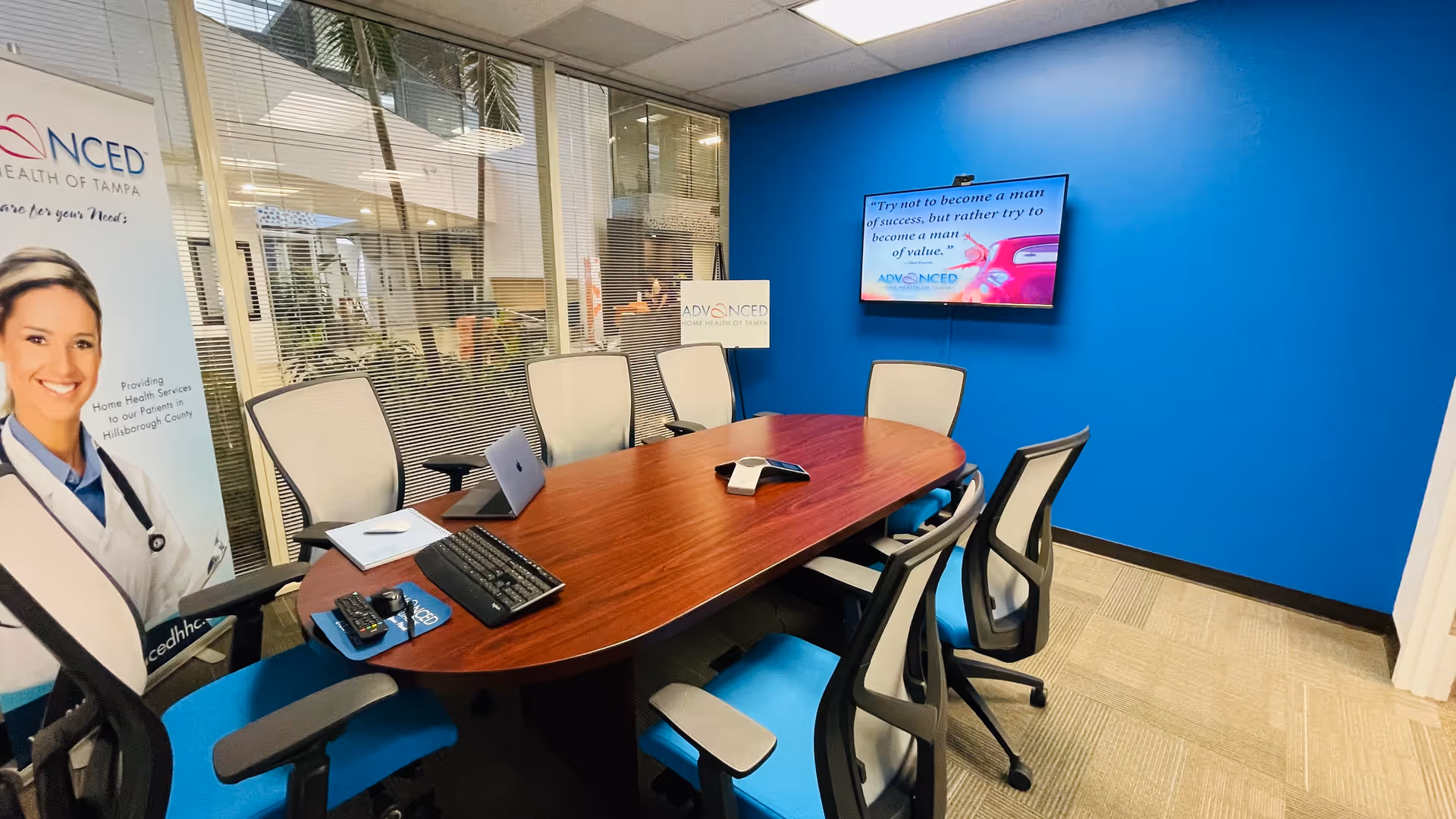 Conference room with wooden table, blue wall, multiple chairs, and a wall-mounted screen.