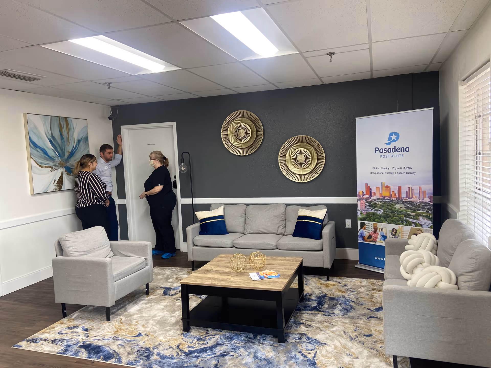A furnished reception-style living room with gray sofas, armchairs, a coffee table, decorative wall art, and three people talking near a door beside a Pasadena Post Acute banner.