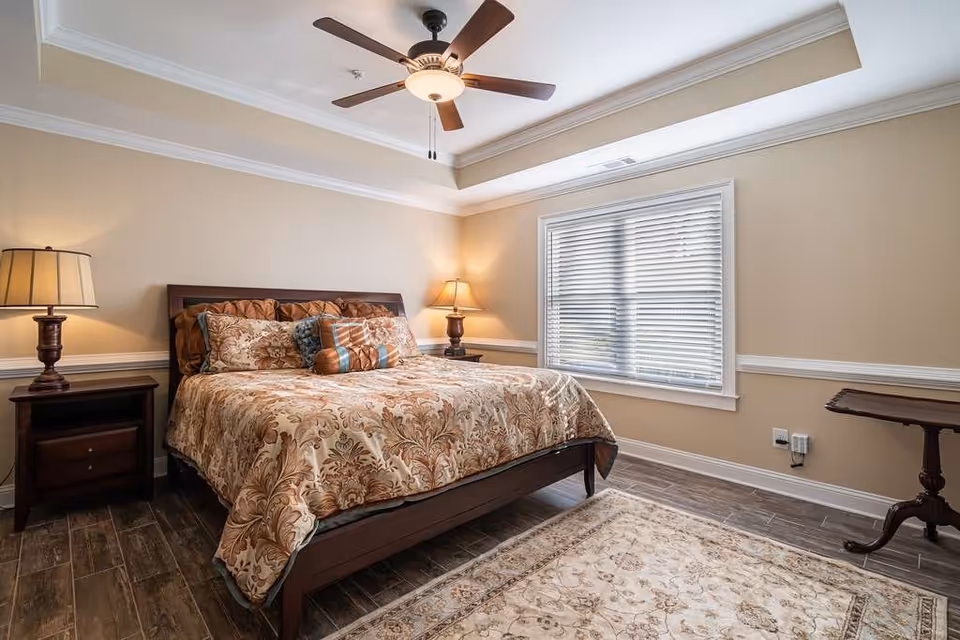 A cozy bedroom with a large bed featuring a patterned bedspread and multiple pillows. There are two wooden nightstands with lamps on either side of the bed, a ceiling fan with a light fixture above, a window with blinds, and a decorative rug on the wooden floor.