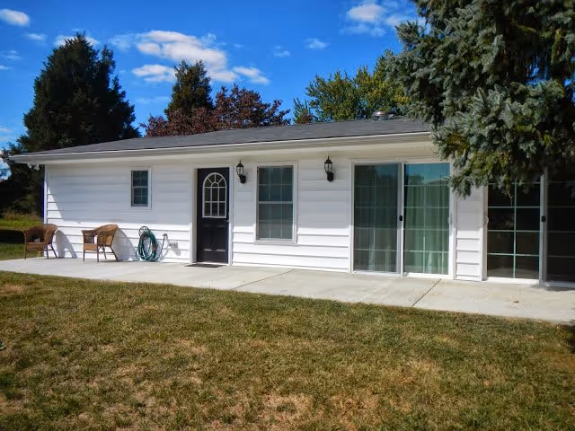 Single-story white building exterior with a front door, windows, chairs, and a concrete path.