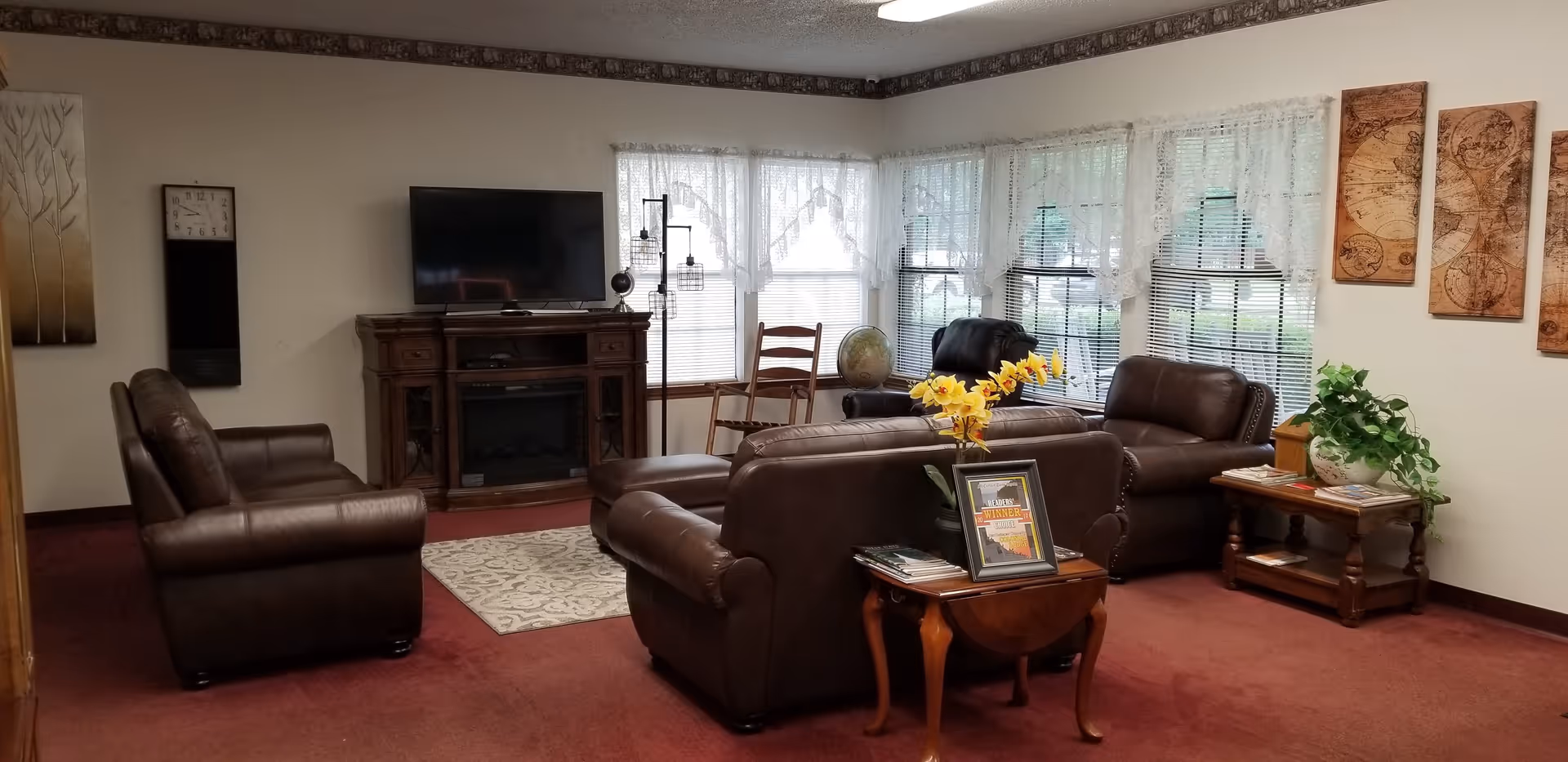 A cozy senior living room with brown leather sofas and armchairs arranged around a wooden TV stand with a flat-screen TV. The room has large windows with white lace curtains, a floor lamp, a globe, and decorative wall art. There are small wooden tables with plants, magazines, and a framed award on one of the tables. The carpet is red, and the walls are light-colored with a decorative border near the ceiling.