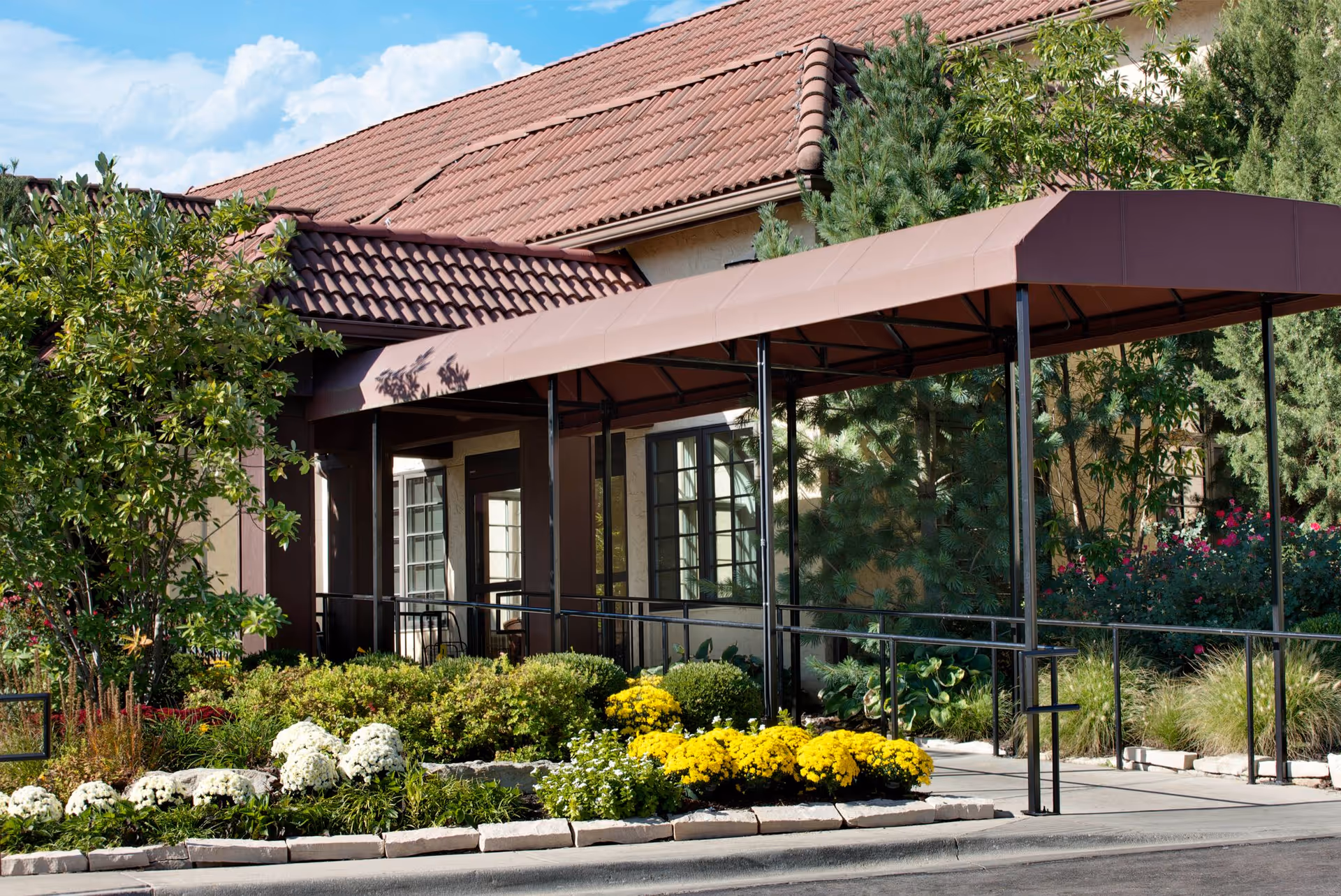 Covered entrance with a metal canopy and accessibility ramp leading to a tile-roofed senior living building surrounded by landscaped flowers and shrubs.