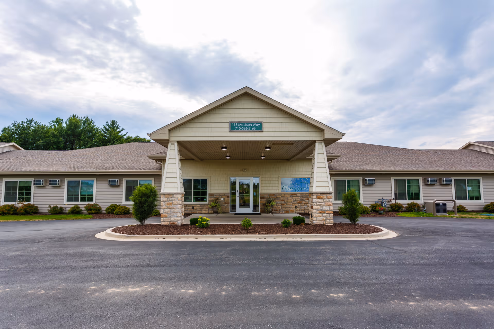Front entrance of a single-story memory care building with a covered drop-off area, central glass doors, and small landscaped islands.