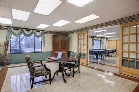 Common dining area with a round table, floral chairs, windows, and an adjoining room.