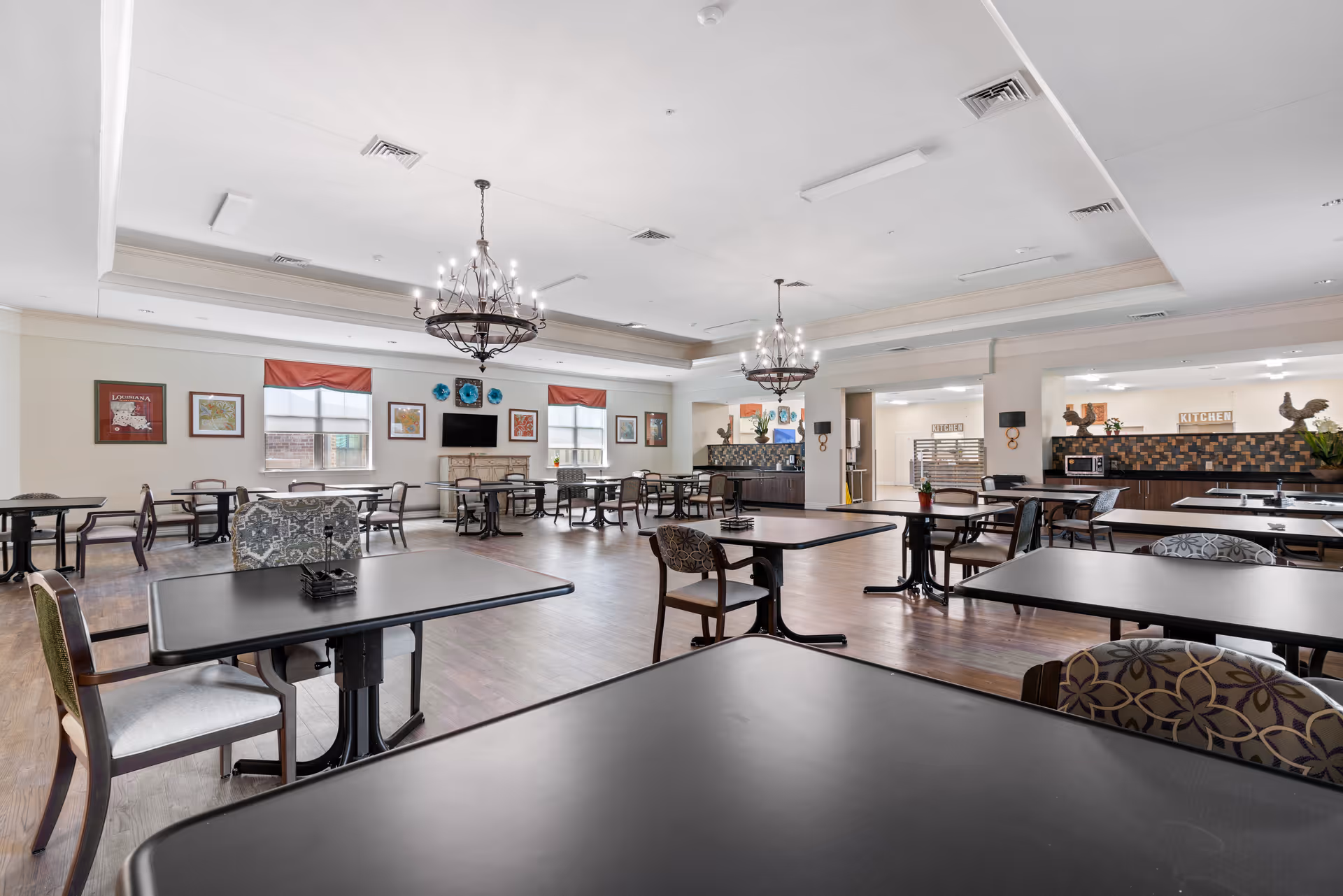 A spacious dining room with multiple black tables and assorted chairs arranged neatly. The room features two large chandeliers hanging from the ceiling, framed artwork on the walls, and windows with red valances allowing natural light to enter. In the background, there is a kitchen area with a tiled backsplash and various kitchen appliances.