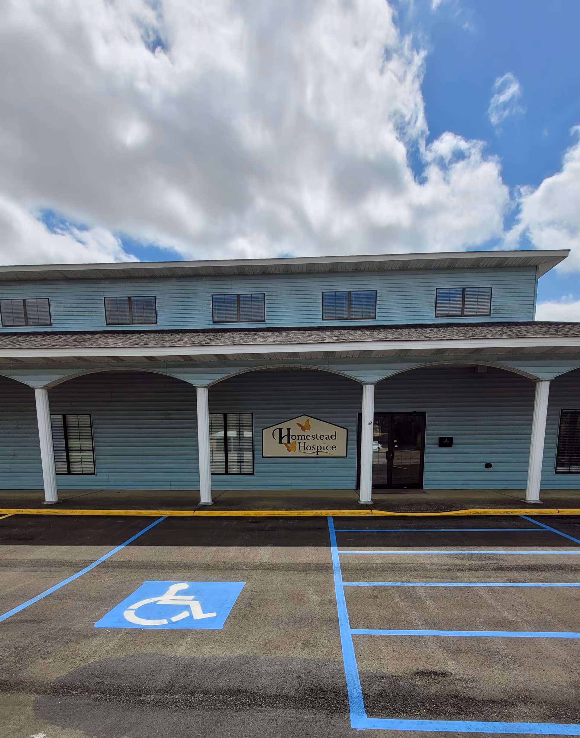 Blue, single-story facility front with arched entrance, sign reading Homestead Hospice, handicap parking.