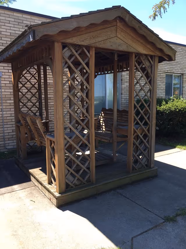 Wooden gazebo with lattice sides and benches sits on a concrete patio outside a brick building.