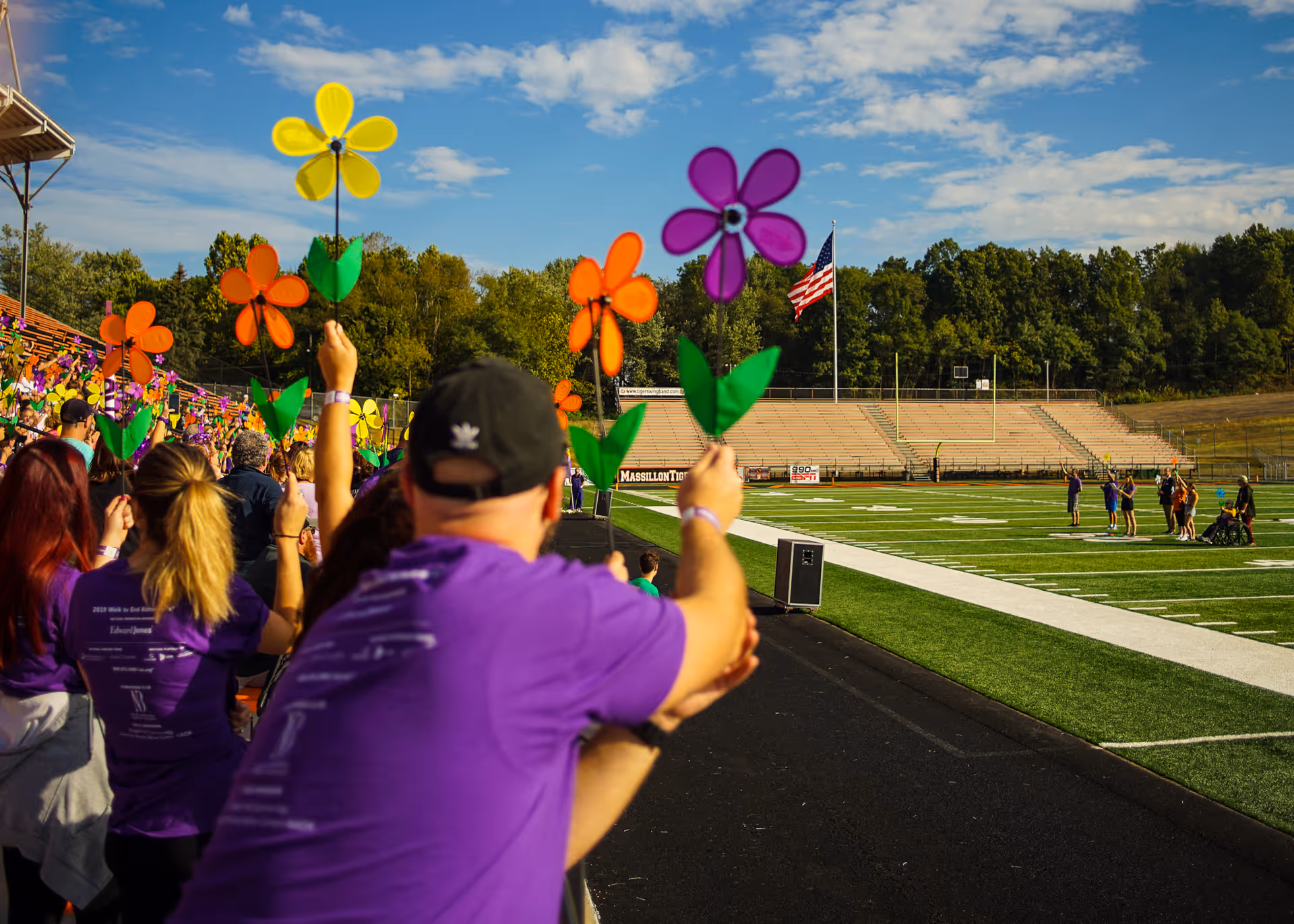 People in purple shirts sitting in stadium bleachers holding colorful pinwheel flowers during a sunny day at a sports field with an American flag in the background.