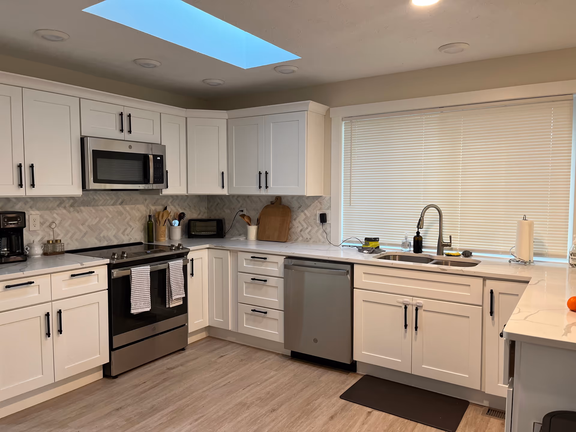 Bright white kitchen with stainless appliances, skylight, and large window with blinds.