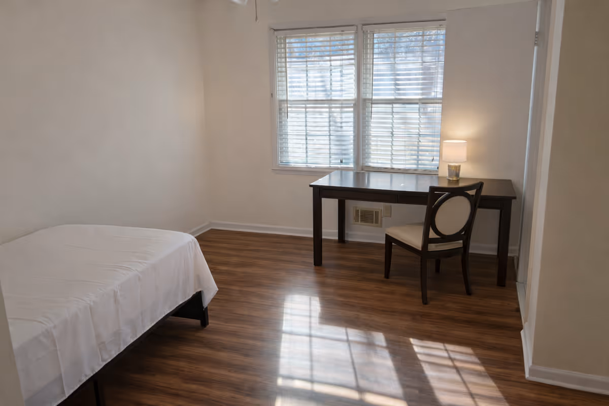 A simple bedroom in a senior care facility featuring a bed, desk, chair, and window.