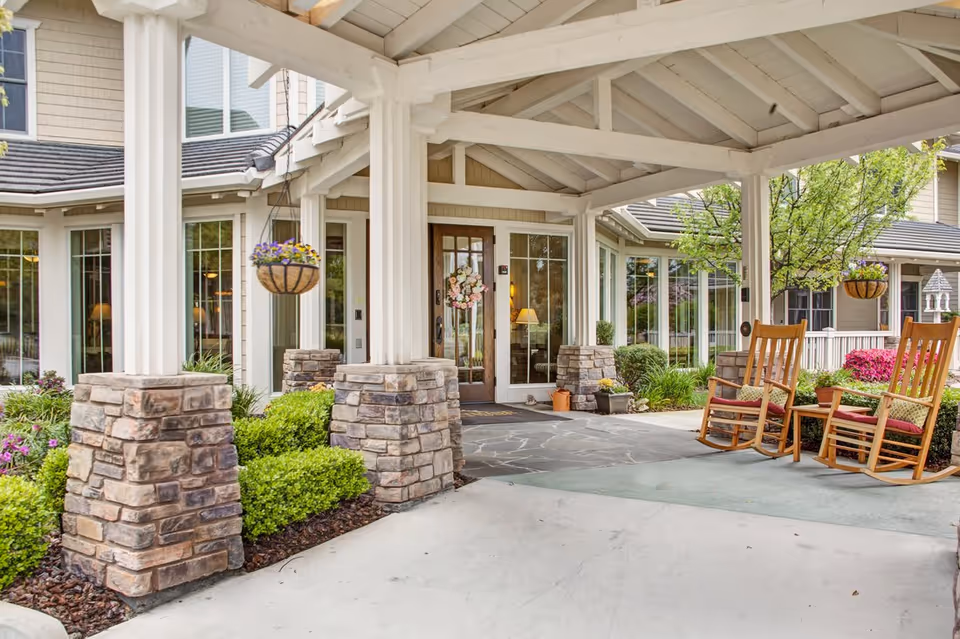 Covered front entrance with stone columns, hanging flower baskets and wooden rocking chairs on a porch.