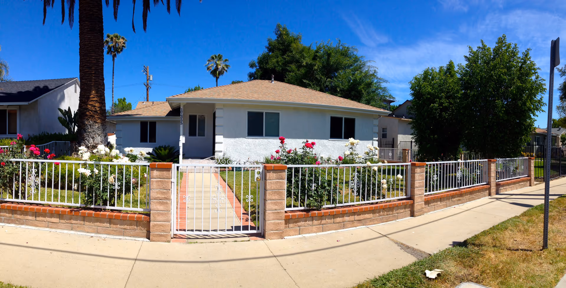 Front exterior of a single-story white stucco house with a gated brick-and-iron fence and rose bushes under a clear blue sky.