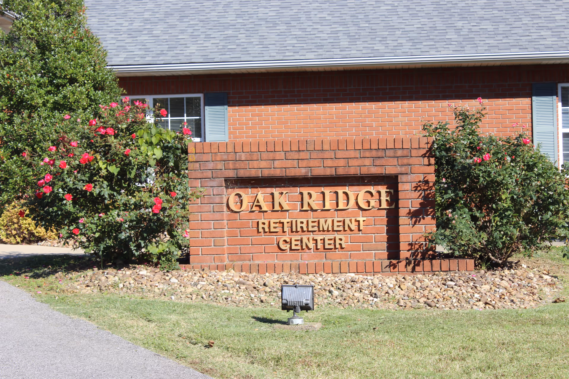 A brick sign in front of a building with the text 'OAK RIDGE RETIREMENT CENTER' surrounded by bushes with pink flowers and a small spotlight in front of the sign.