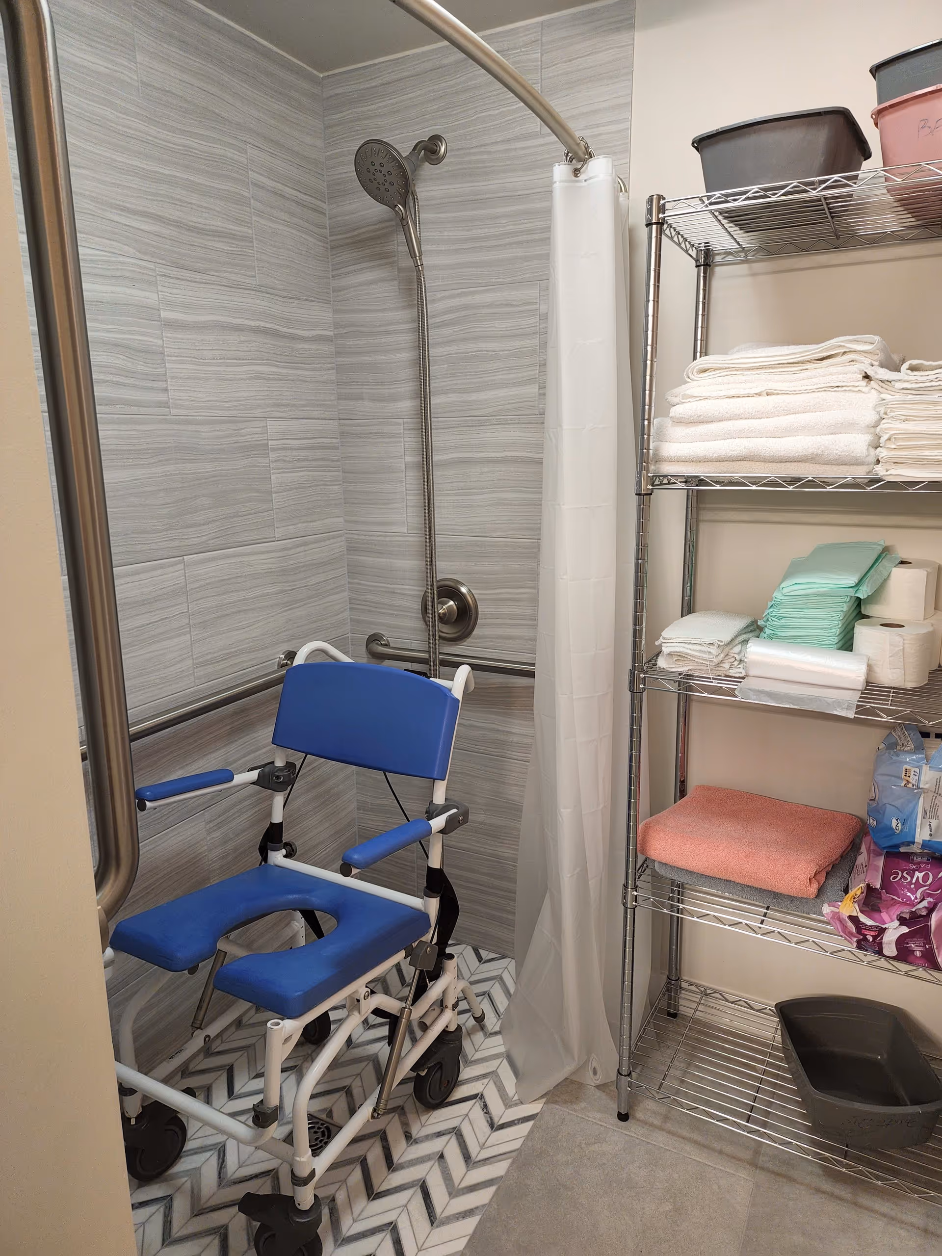 A bathroom shower area with a blue shower chair on wheels, a handheld showerhead mounted on a tiled wall, and a white shower curtain. Next to the shower is a metal shelving unit stocked with folded white towels, green disposable pads, toilet paper rolls, a pink towel, and other supplies.