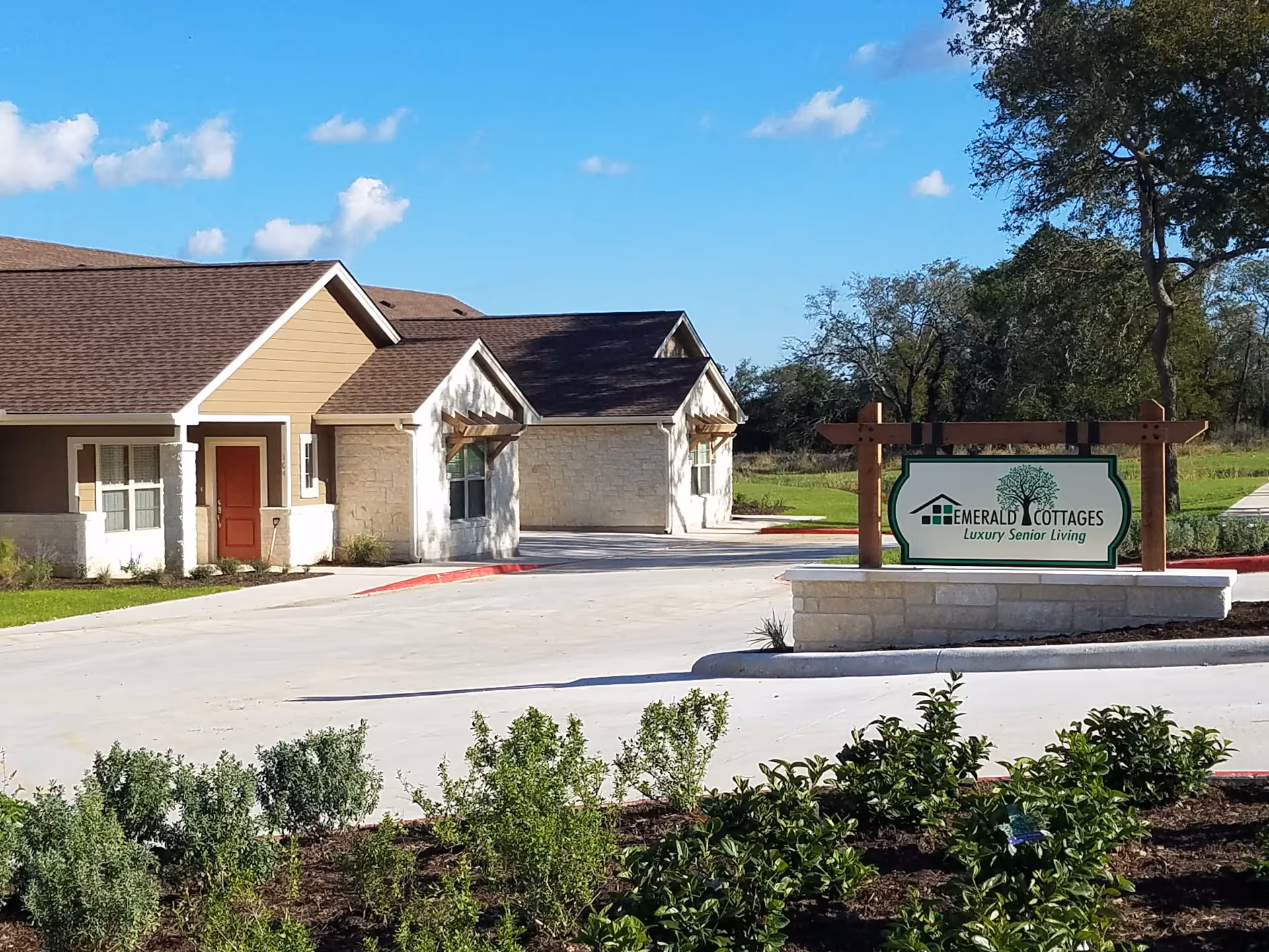 Exterior view of a senior living facility named Emerald Cottages of Kerrville, showing a driveway, landscaped bushes, and a sign that reads 'Emerald Cottages Luxury Senior Living' with a tree logo under a clear blue sky.