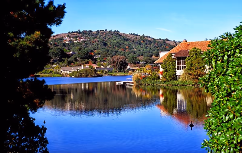 A tranquil lakeside scene with a building, trees, hills, and reflections.