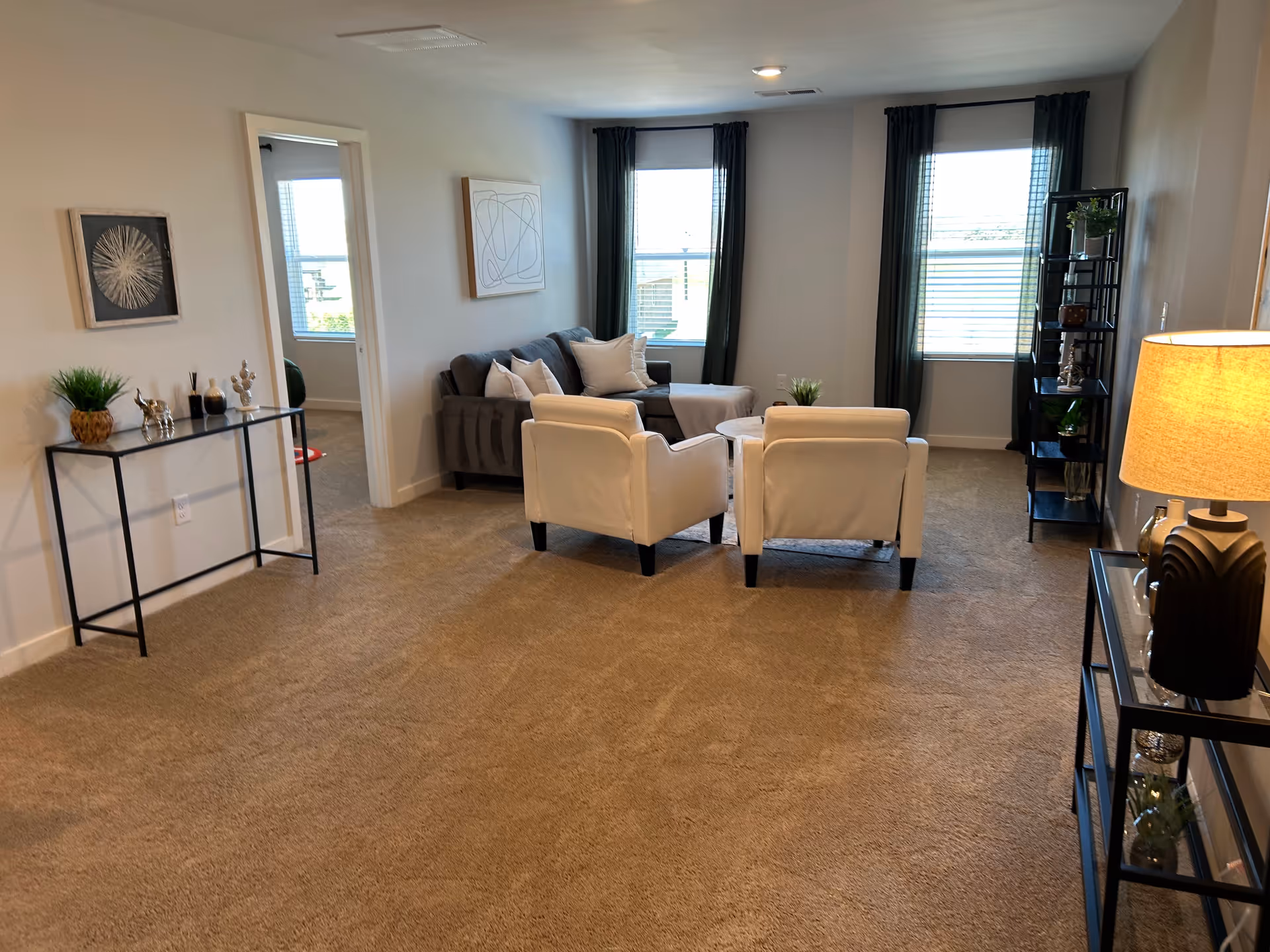 Bright living room with beige carpet, sofa, chairs, and large windows in a senior facility.