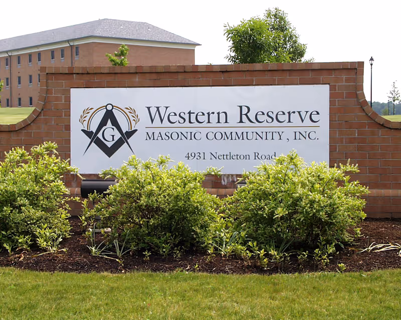 A brick sign with a white panel displaying the text 'Western Reserve Masonic Community, Inc. 4931 Nettleton Road' along with a Masonic symbol. The sign is surrounded by green bushes and grass, with a building and trees visible in the background.