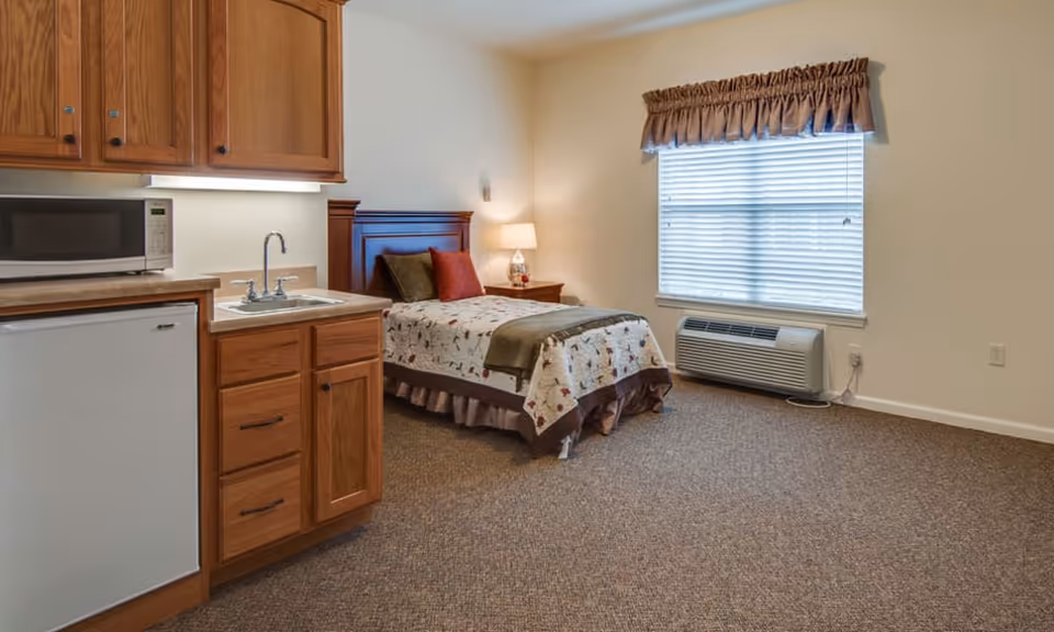 A studio-style senior living bedroom with a single bed, bedside table and lamp, a small kitchenette with sink and microwave, and a window air conditioner.