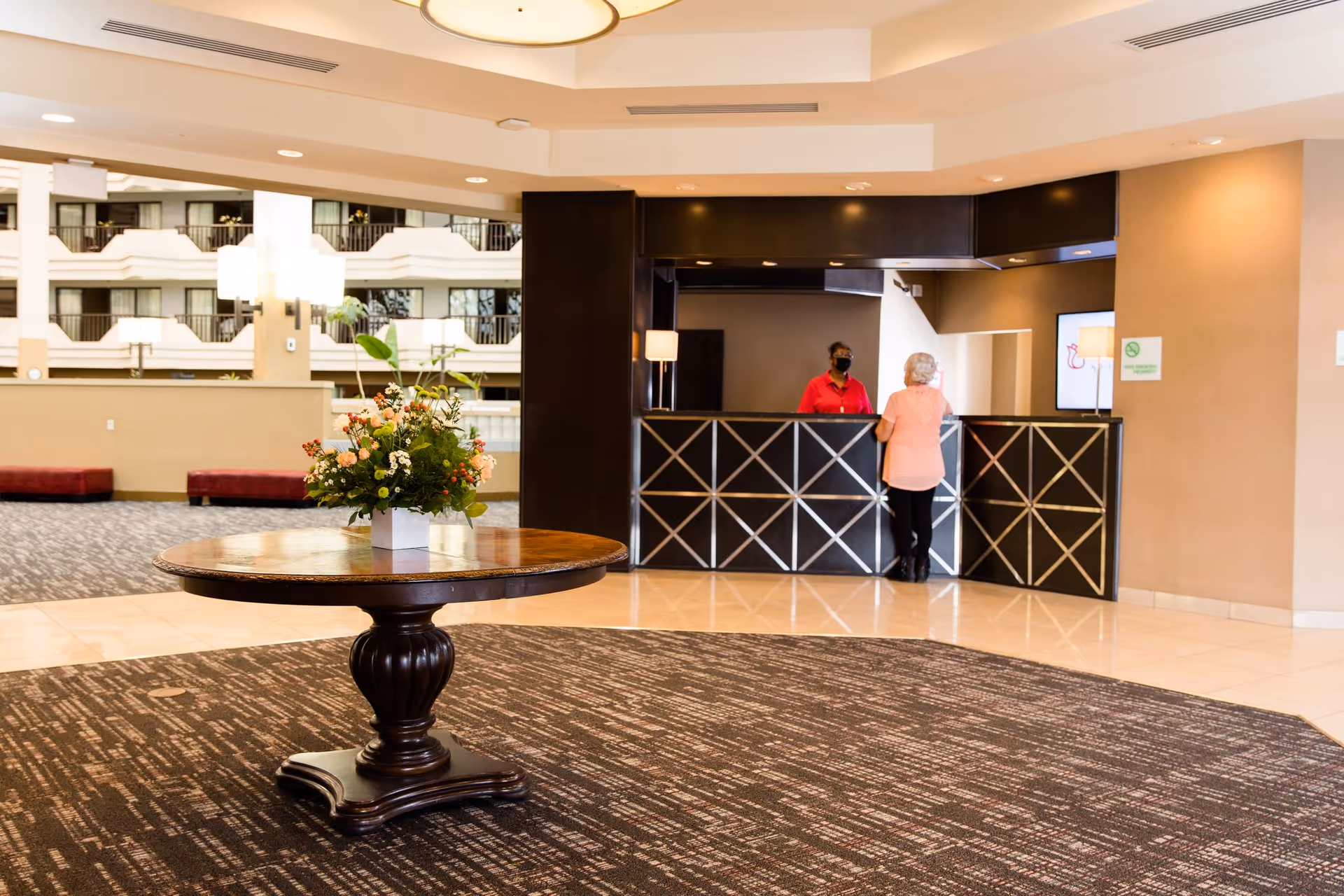 A spacious assisted living facility lobby with a round wooden table holding a floral arrangement in the foreground. In the background, a receptionist wearing a red shirt and black face mask stands behind a dark reception desk, speaking with an elderly woman in a peach top. The area features beige walls, patterned carpet, and a large open atrium with balconies visible.