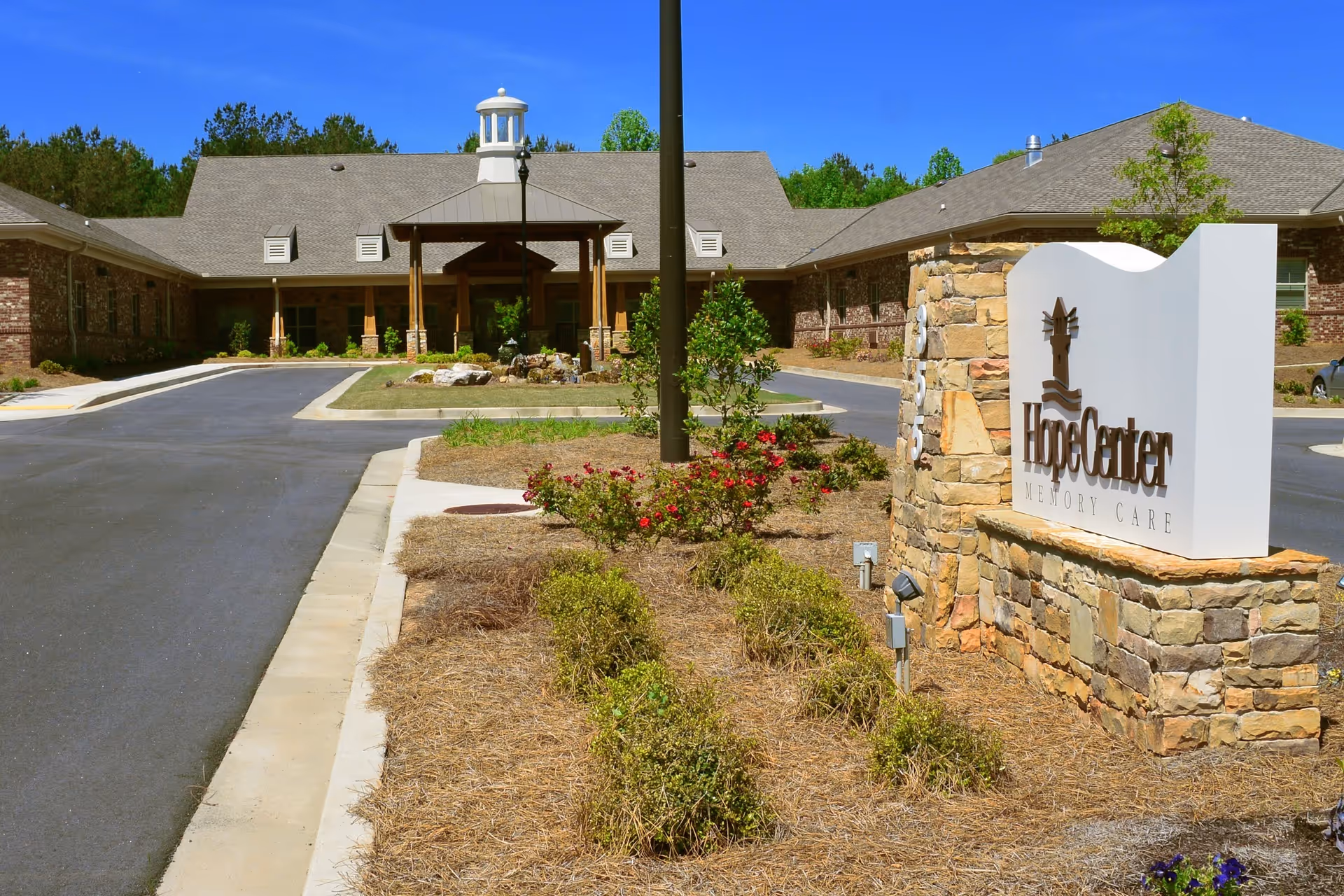 Exterior view of Hope Center Memory Care facility showing a single-story brick building with a covered entrance, a driveway, landscaped bushes, and a stone sign with the facility's name.