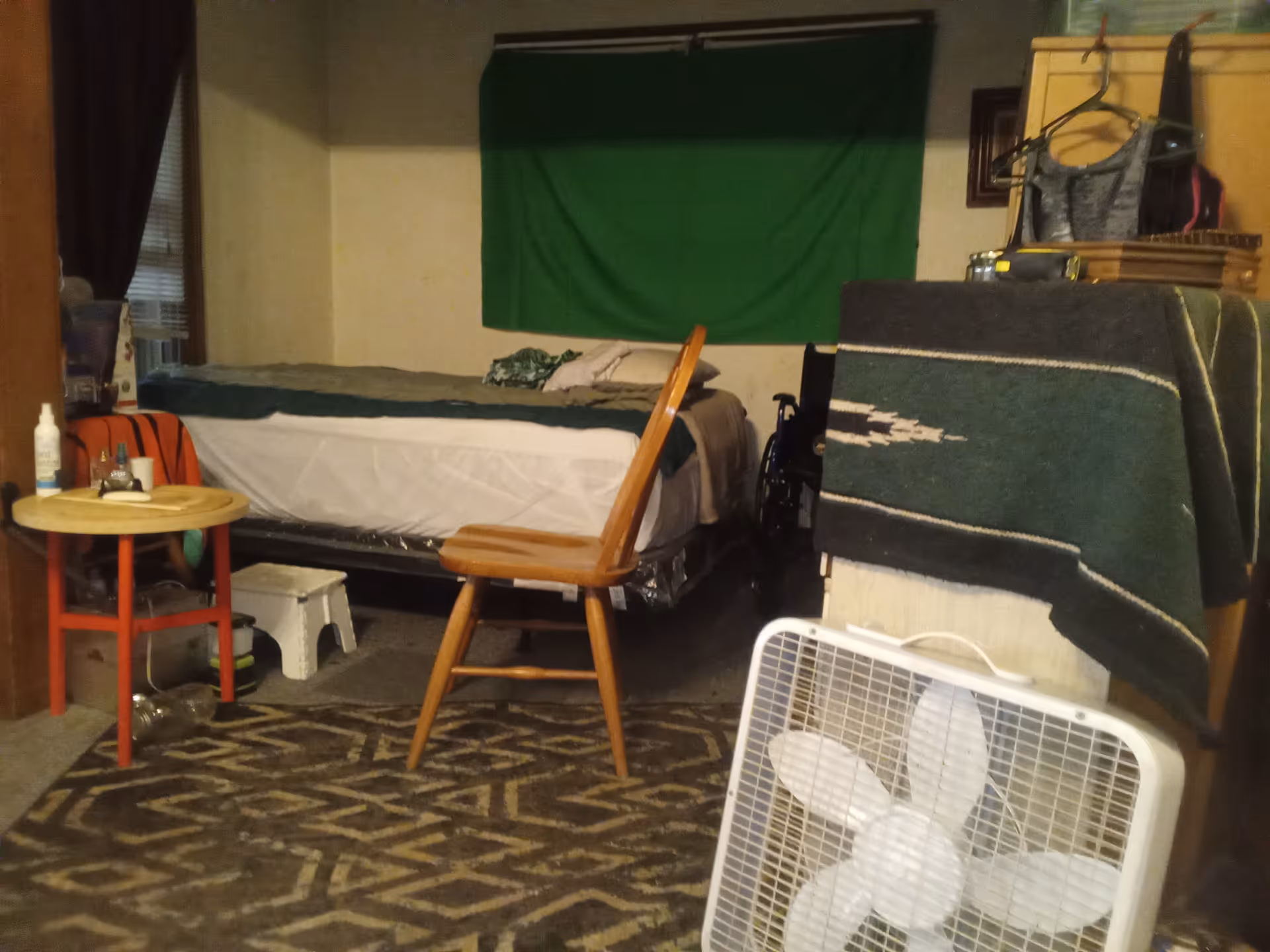 A cluttered bedroom in a senior facility with bed, chair, table, and standing fan.