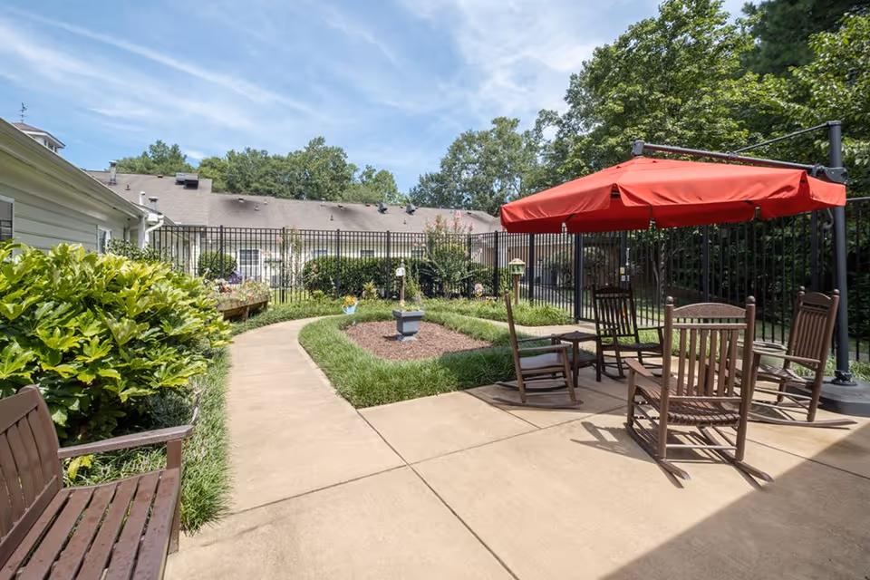 Sunny fenced courtyard with a paved walkway, garden beds, wooden rocking chairs and a red patio umbrella surrounded by shrubbery.