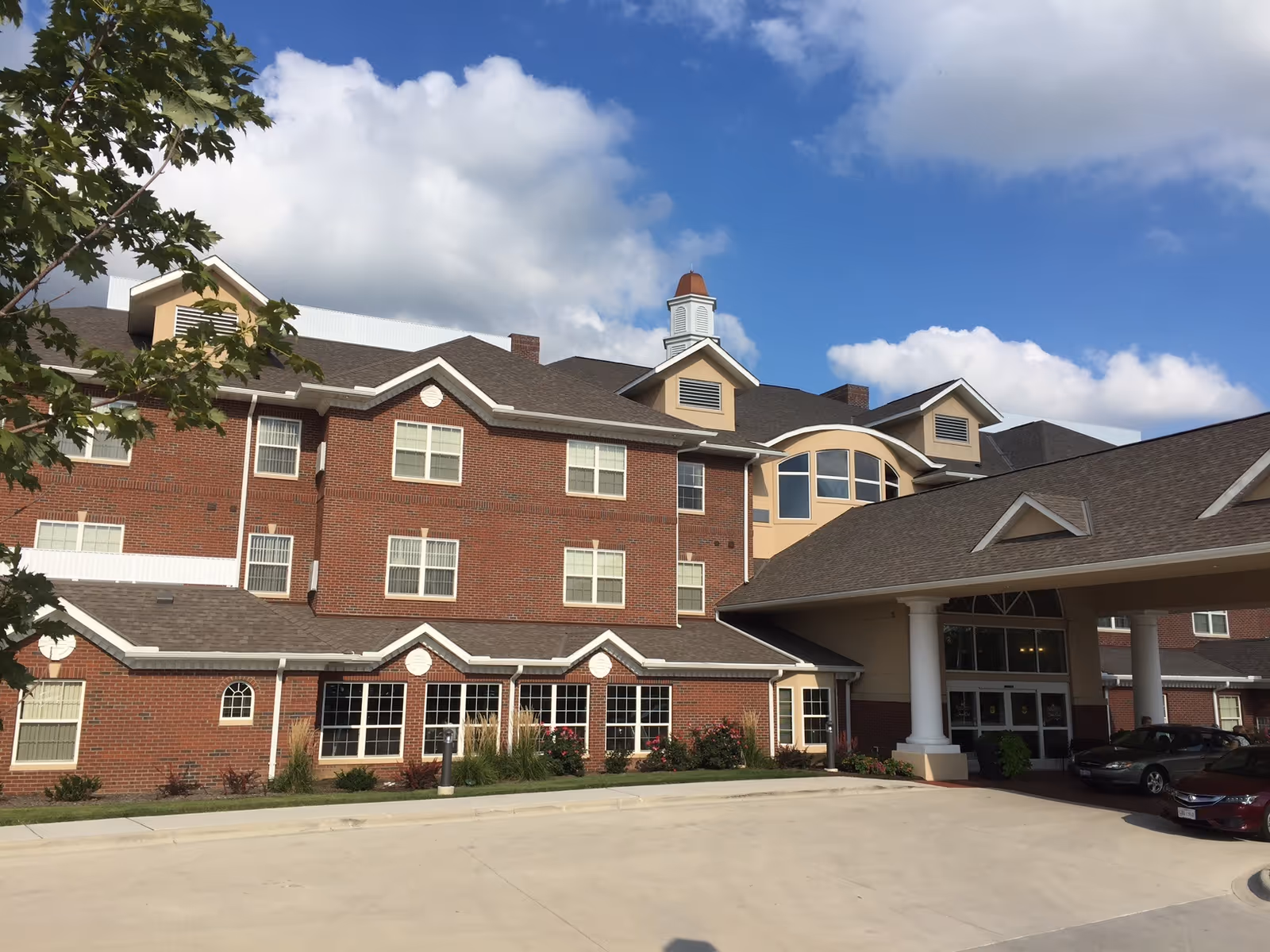 Front exterior of a multi-story red brick senior living building with a covered entrance and parked cars under a blue sky.