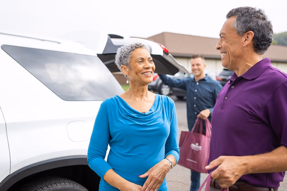 An older woman in a blue top and an older man in a purple polo shirt smiling and talking near a white SUV with its trunk open. In the background, a younger man is holding a maroon bag and standing near the vehicle in a parking lot outside a building.