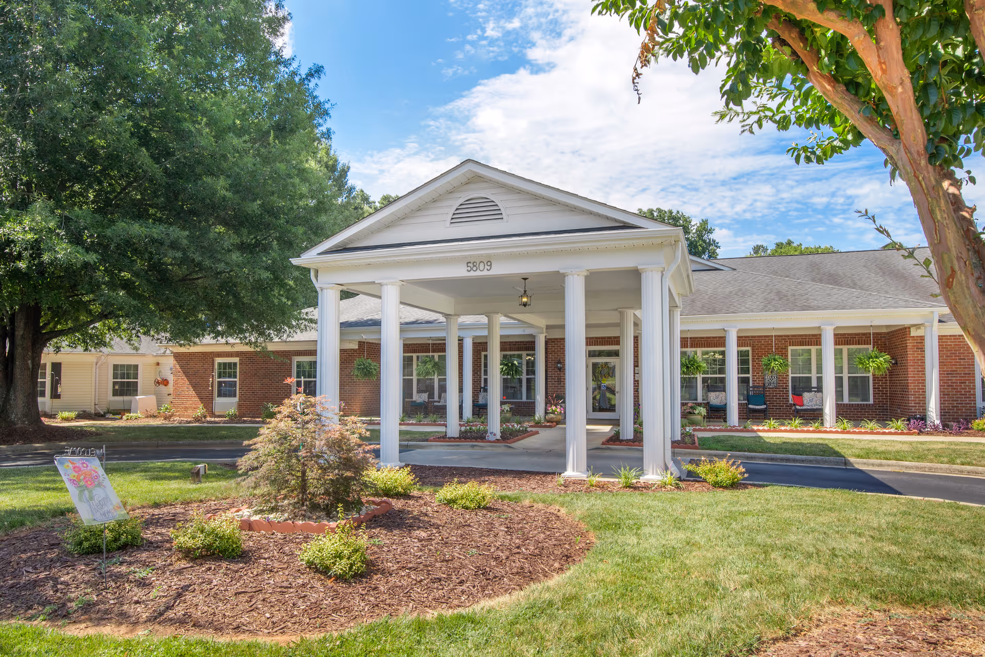 Front exterior view of Brookdale Northwest Greensboro facility featuring a covered entrance with white columns, brick walls, hanging plants, and a landscaped garden with green grass and small shrubs under a partly cloudy blue sky.