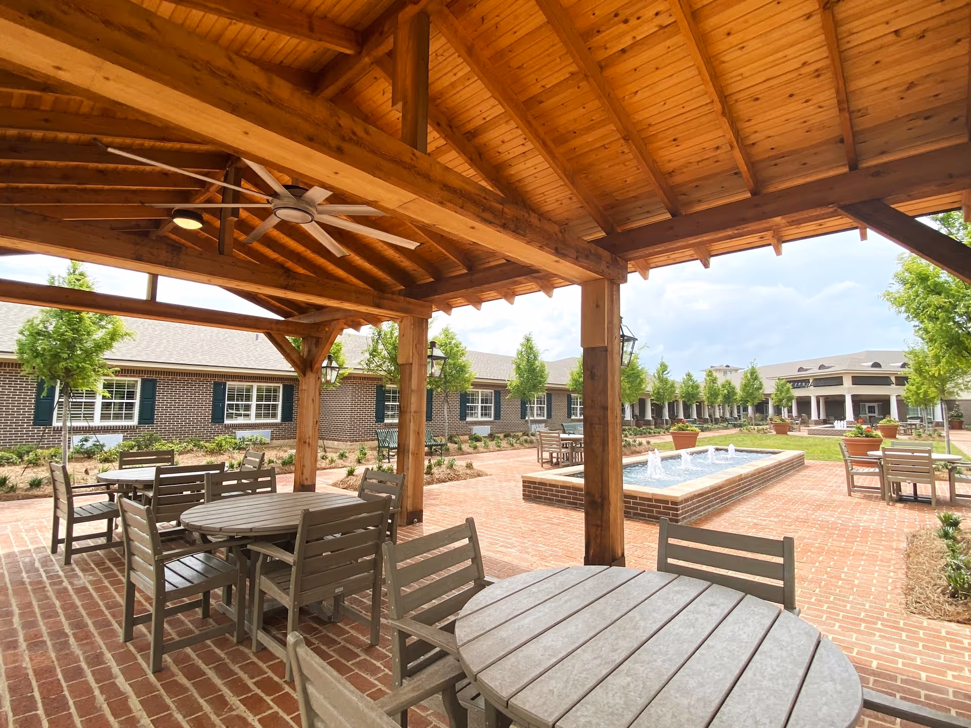 Outdoor covered seating area with round tables and chairs under a wooden pergola with a ceiling fan. In the background, there is a brick-paved courtyard with a rectangular water fountain, trees, and a large building with multiple windows and green shutters.