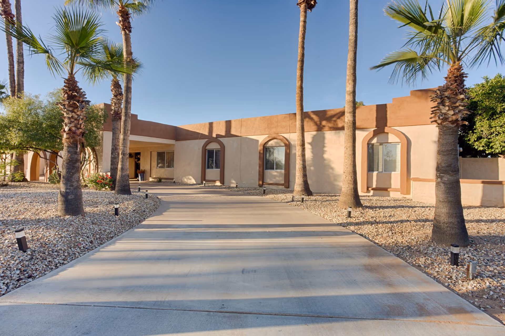 Front exterior of a single-story stucco building with arched windows, a driveway, and palm trees.