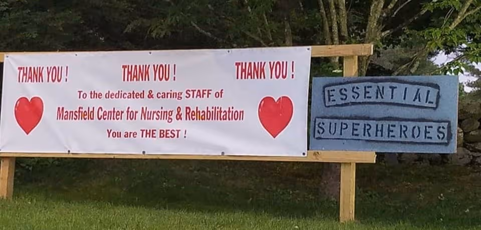 Outdoor banner thanking Mansfield Center for Nursing and Rehabilitation staff, next to blue 'Essential Superheroes' sign.