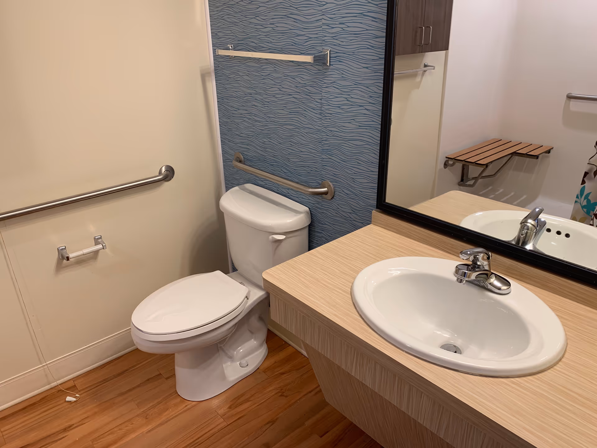 A bathroom with a white toilet and a sink with a silver faucet on a wooden countertop. The wall behind the toilet has a blue textured design and a horizontal grab bar. There is a large mirror above the sink, a towel bar, and a foldable wooden shower seat mounted on the wall. The floor is wooden.